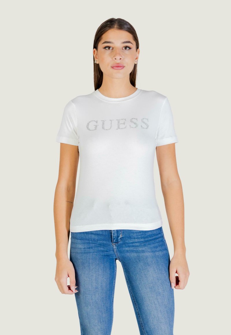 T-shirt bianca a maniche corte con logo "GUESS" in argento sul petto, realizzata in tessuto liscio. Abbinata a jeans blu.