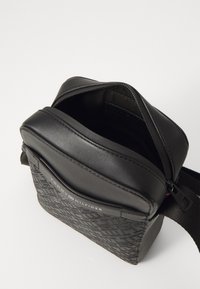 Svart crossbody-veske med et teksturert nylonmateriale, lærdetaljer og glidelås. Har et logo-patch og justerbar stropp.