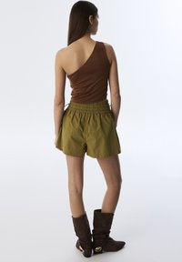 Haut marron à une �épaule associé à un short vert olive avec une taille élastique et une coupe ample, complété par des bottes marron jusqu'aux genoux.