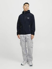 Marineblauwe softshell jas met capuchon, ritszakken en logodetail. Gepaarde met lichtgrijze cargobroek en sportschoenen.