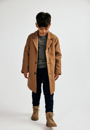 Kreemivärviline villane mantel, millel on lõhiku krae, hall särk all, tumesinised püksid ja kreemivärvilised saapad. Mantel on varustatud nööpidega ja sellel on mugav lõige.