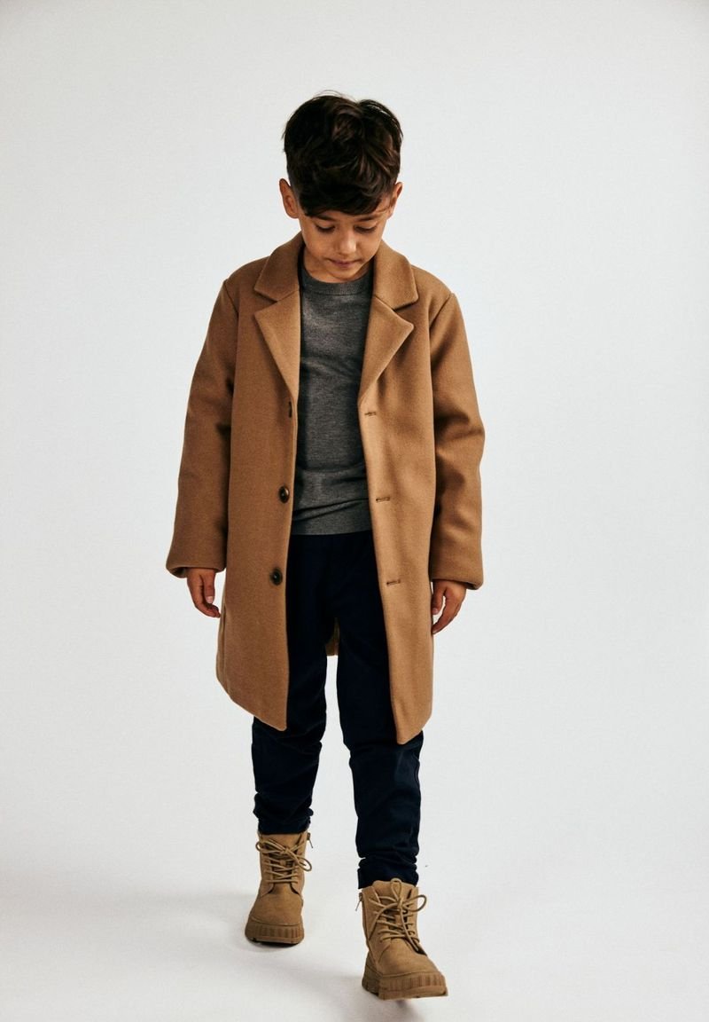 Kreemivärviline villane mantel, millel on lõhiku krae, hall särk all, tumesinised püksid ja kreemivärvilised saapad. Mantel on varustatud nööpidega ja sellel on mugav lõige.