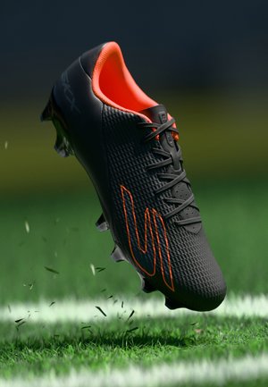 Čierna futbalová topánka s textúrovaným povrchom, výraznými oranžovými prvkami a šnurovaním, zobrazujúca kopačky a zdvihnuté trávové listy.