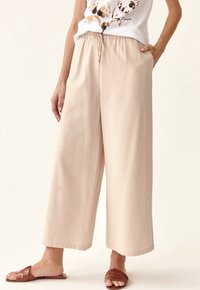 Pantalons beige à jambes larges avec taille à cordon et poches latérales, fabriqués en tissu lisse. Portés avec des sandales brunes à brides.