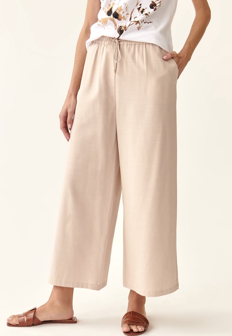 Pantalons beige à jambes larges avec taille à cordon et poches latérales, fabriqués en tissu lisse. Portés avec des sandales brunes à brides.