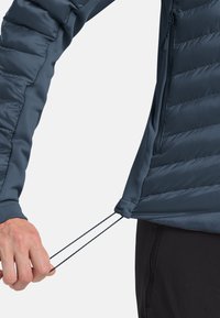 Tirer à la main sur le cordon de serrage à l'ourlet d'une doudoune bleue portée sur un pantalon noir, mettant en avant la fonctionnalité d'ajustement.