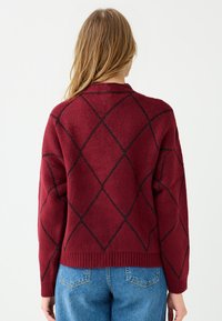 Kobieta z blond włosami, ubrana w czerwony sweter w czarny wzór w romby i niebieskie dżinsy, pokazana plecami na białym tle.