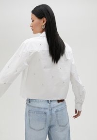 Chemise blanche raccourcie avec des détails en perles argentées, manches longues et col classique, associée à un jean bleu clair.