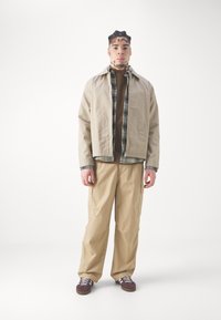 Beige bomuldsjakke over en brun sweater, parret med khaki cargo bukser og maroon sko med hvide snørebånd. Ternet detaljer på jakken.
