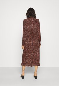 Robe longue à fleurs avec une base rouge foncé ornée de motifs blancs et bleus, fabriquée en tissu léger. Elle a des manches longues et un ourlet fluide.