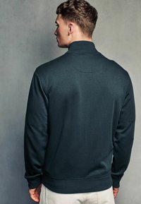 Sweat-shirt vert foncé avec un col montant, texture lisse et poignets côtelés. Le dos présente un détail de couture circulaire.