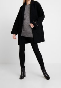 Black oversized coat with a textured finish, featuring two front pockets and a single button closure, worn over a metallic gray top and black pants.