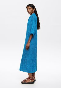 Abito lungo in maglia color teal con motivi intricati e trame aperte. Presenta un colletto e maniche fluide. Abbinato a sandali marroni con lacci.