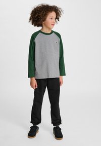 Grijs langarm T-shirt met groene raglanmouwen en een klein embleem op de borst, gecombineerd met zwarte trainingsbroek en zwarte schoenen.