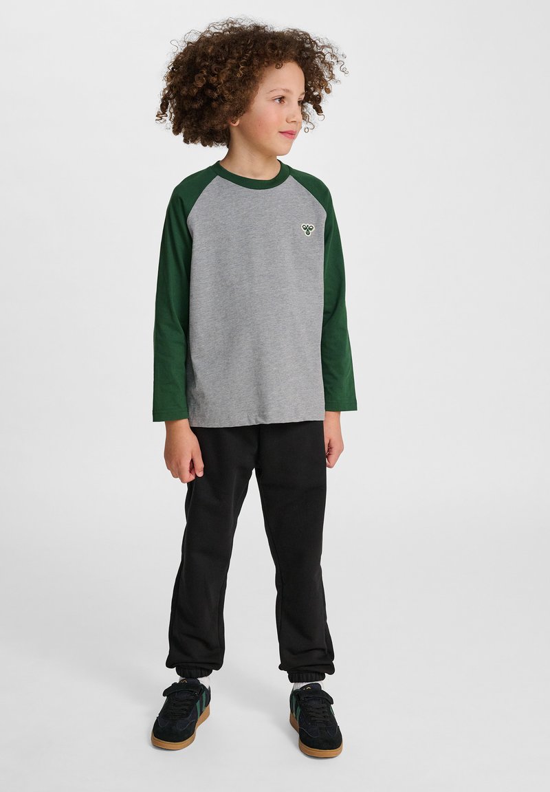 Grijs langarm T-shirt met groene raglanmouwen en een klein embleem op de borst, gecombineerd met zwarte trainingsbroek en zwarte schoenen.