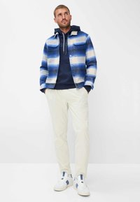 Rutet jakke i blå og hvid, marineblå hoodie under, cremefarvede kordfløjspants og hvide sneakers med blå accenter.