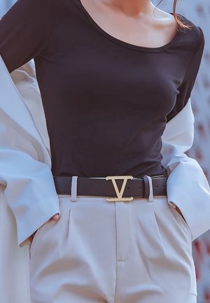 Vrouw gekleed in een zwart topje met een ronde hals, beige hoge taillebroek met zwarte riem met gouden "V" gesp, en een lichtbeige jasje over haar schouders gedrapeerd.