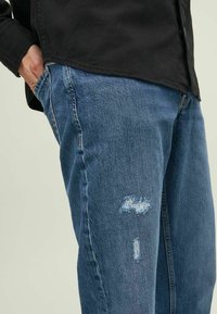 Vaqueros de denim azul con un corte relajado y detalles desgastados, incluyendo pequeños desgarros en el muslo y costuras contrastantes.