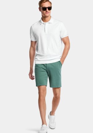 Jeune homme portant des lunettes de soleil noires, un polo blanc, un short vert et des baskets blanches, marchant avec une main dans la poche.