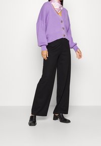 Lila gebreide cardigan met oversized mouwen en drie grote knopen, gecombineerd met zwarte wijde broek en zwarte blokhak schoenen.
