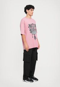 Άνδρας που φοράει ροζ υπερμεγέθη T-shirt με σχέδιο κλουβιού πουλιών, μαύρο cargo παντελόνι με πολλές τσέπες και μαύρα sneakers με λευκές σόλες.