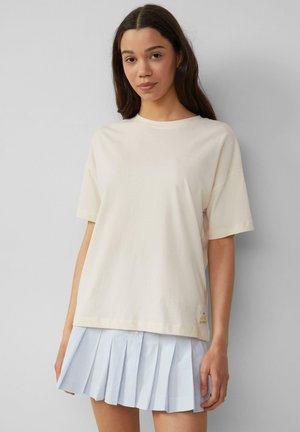 T-shirt en coton crème à manches courtes et col rond. Présente une petite étiquette graphique en bas. Associé à une jupe plissée bleu clair.