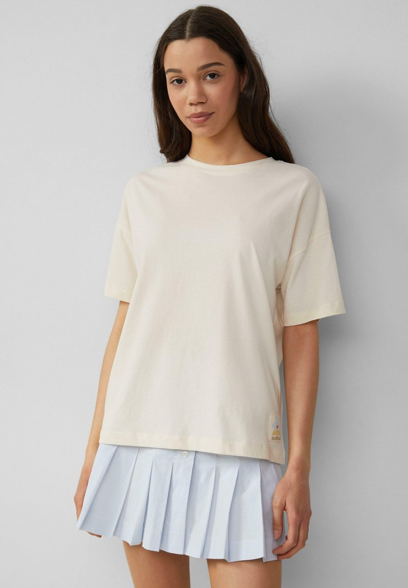 T-shirt en coton crème à manches courtes et col rond. Présente une petite étiquette graphique en bas. Associé à une jupe plissée bleu clair.