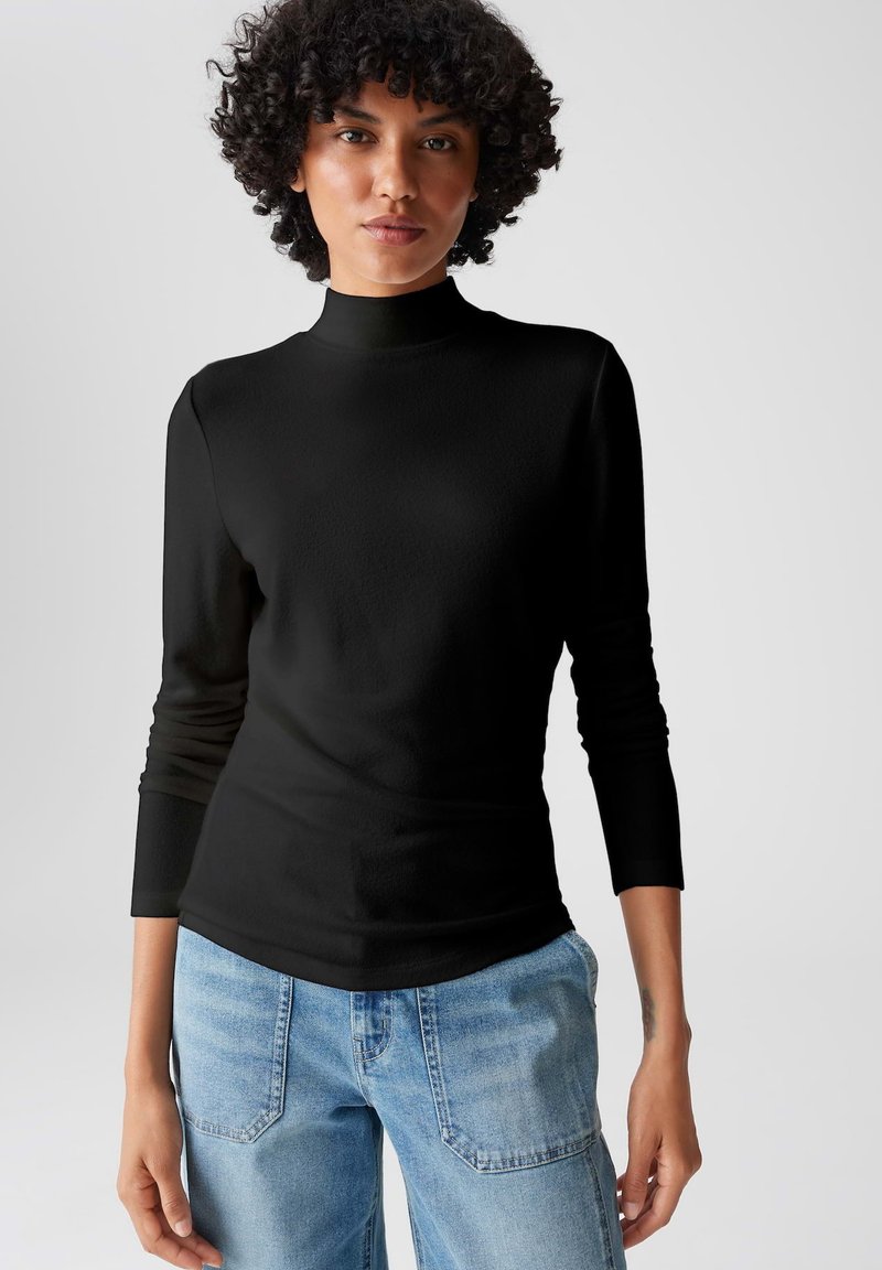 Schwarzes langärmliges Rollkragenoberteil mit glatter Textur, tailliertem Schnitt und hohem Kragen. Kombiniert mit hellblauen Jeans.