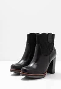 Zwarte enkelboots hebben een glanzend leren voorzijde en een gestructureerde suède bovenkant, met een hoge blokhak en gestikte accenten op de zool.