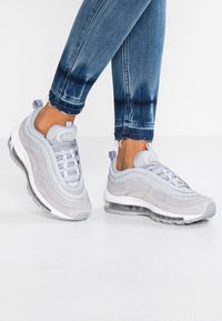 Grå sportskor med ett lagerdesign, silveraccenter, överdel av mesh och en synlig luftdämpande sula, tillsammans med slitna blåjeans.