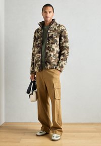 Jakna od kamuflaže od flisa s visokim ovratnikom, uparena s khaki cargo hlačama. Outfit uključuje sive i bijele tenisice te malu torbu.