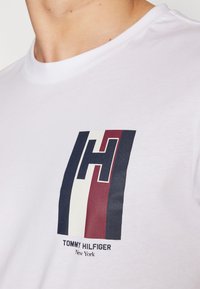 Bílá bavlněná tričko s grafickým potiskem s velkým "H" v námořnické modré, červené a bílé pruhy. Pod logem je text "TOMMY HILFIGER New York."