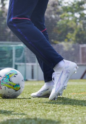 Baltas futbols ar glancētu virsmu un varavīksnes akcentiem, apvienots ar tumši ziliem biksēm, novietots virs krāsaina futbolbala uz zāliena.