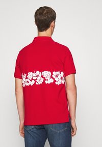 Polo rouge avec un motif floral blanc au dos, à manches courtes et col. Le tissu semble lisse avec des fentes sur les côtés à l'ourlet.