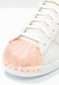 Vit mockaskor med en glänsande roséguldshelltoe, med upphöjda linjer och en vit gummisula, som visar en texturerad yta.