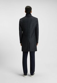 Cappotto blend di lana grigio scuro con colletto alto, caratterizzato da un motivo testurizzato e un taglio dritto, abbinato a pantaloni blu navy e sneakers chiare.