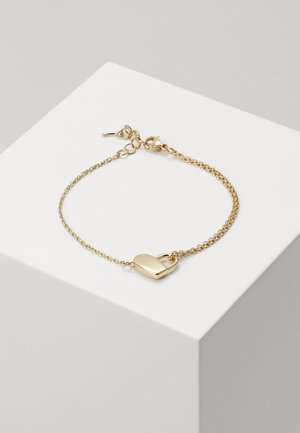 Delikatna złota bransoletka z łańcuszkiem z małym wisiorkiem w kształcie serca, prezentowana na białej geometrycznej powierzchni.