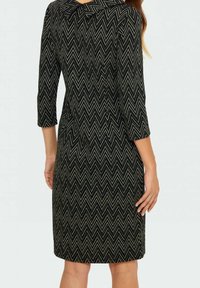 Černé šaty ke kolenům s bílým cik-cak vzorem a rukávy 3/4, zobrazené ze zadní strany na stojící osobě.