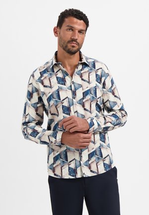 Man in een overhemd met lange mouwen, bedrukt met een geometrisch patroon in blauw, wit en bruin, staande met zijn handen gevouwen.
