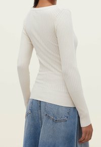 Vrouw in een aansluitende ribben crème lange mouwen top, gecombineerd met losse blauwe denim jeans, gezien vanuit de achterkant.