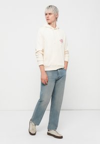 Cream hoodie with a front pocket, red text graphic, paired with light blue straight-leg jeans and beige low-top sneakers.