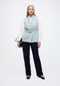 Vestă bleu deschis croită, cu cordon în talie, asortată cu o cămașă albă și pantaloni evazați din denim închis. Modelul ține o geantă neagră și poartă balerini negri.