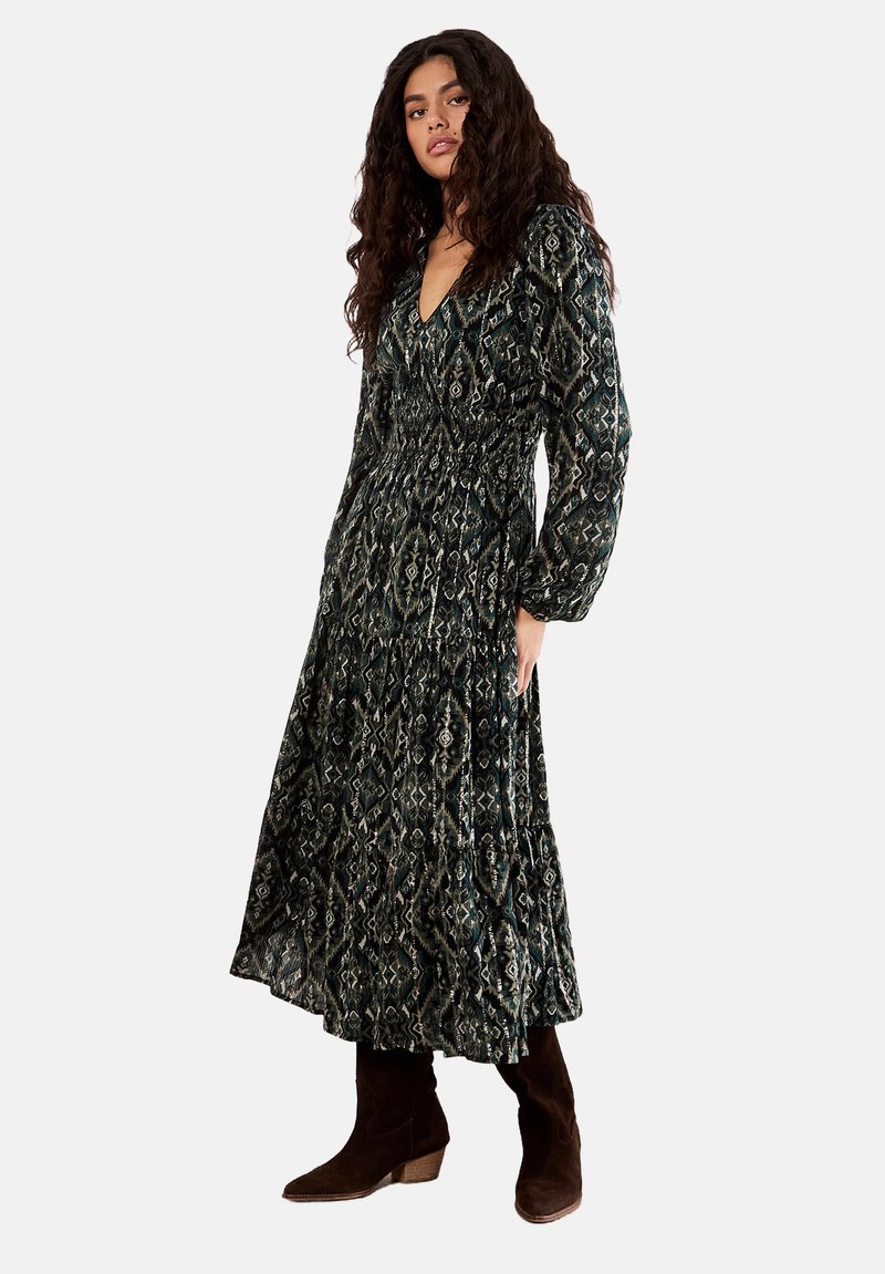 Long dark patterned dress with a V-neck, smocked waist, and billowy sleeves. Paired with brown suede ankle boots. Floral geometric design.