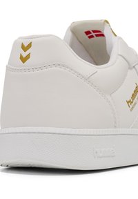 White sneaker with textured leather and rubber sole, featuring gold accents and a chevron design on the heel. Brand name embossed on side.
