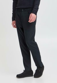 Schwarze, taillierte Hose im Slim-Fit-Schnitt mit elastischem Bund, Knopfverschluss und seitlichen Taschen. Kombiniert mit dunklen Wildlederschuhen.