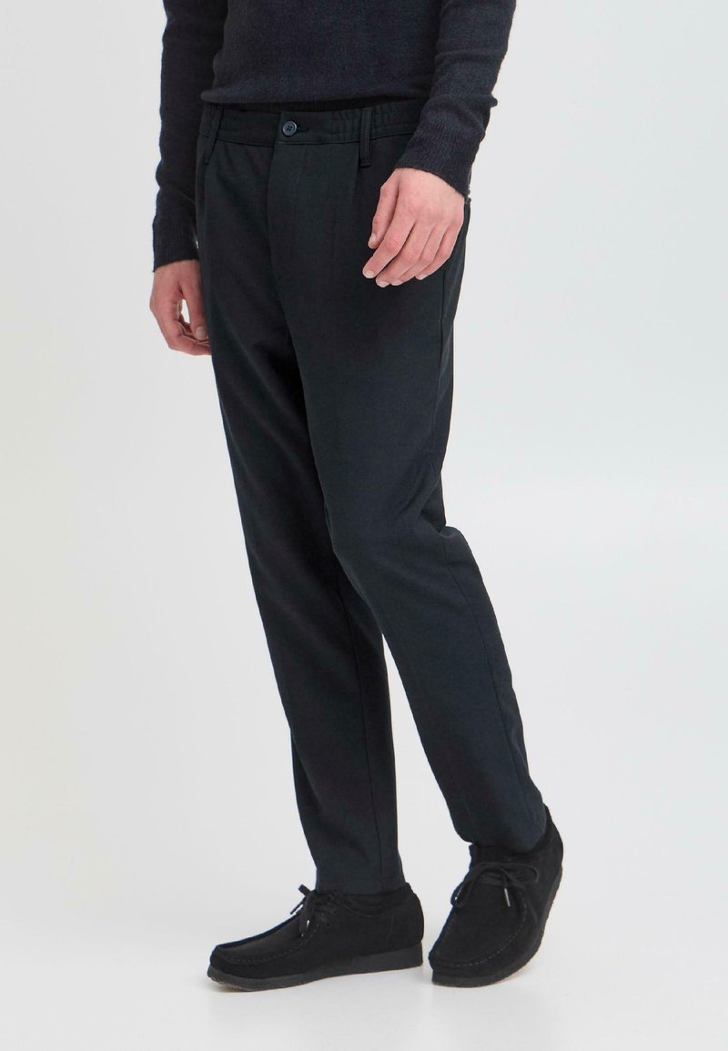 Schwarze, taillierte Hose im Slim-Fit-Schnitt mit elastischem Bund, Knopfverschluss und seitlichen Taschen. Kombiniert mit dunklen Wildlederschuhen.