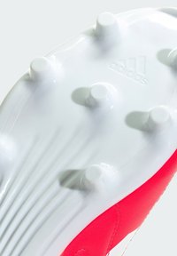 Botas de fútbol con una suela blanca y tacos elevados, y una parte superior sintética de un rojo vibrante, que muestra detalles texturizados y el logo de la marca.