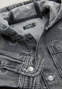 Veste en jean grise avec capuche, présentant des accents de motifs subtils, des coutures audacieuses et des boutons-pression métalliques. L'étiquette indique "NEXT."