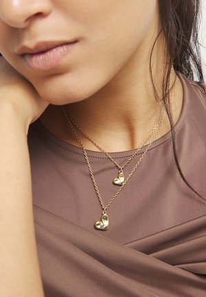 Primer plano del cuello de una mujer que lleva dos colgantes dorados en forma de corazón sobre cadenas de oro en capas, combinados con una blusa marrón.