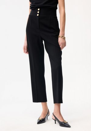 Kobieta nosząca czarne spodenki z wysokim stanem i trzema złotymi guzikami, czarne szpilki na pasku z tyłu oraz złota bransoletka łańcuchowa na białym tle.